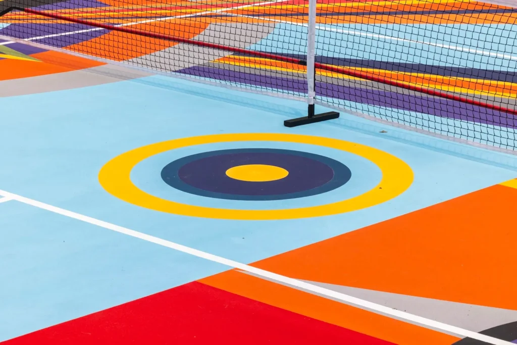 Close-up of an outdoor sports court featuring a large concentric circle target graphic in yellow, navy, and light blue beneath a net, surrounded by bold orange, red, gray, and blue geometric court markings