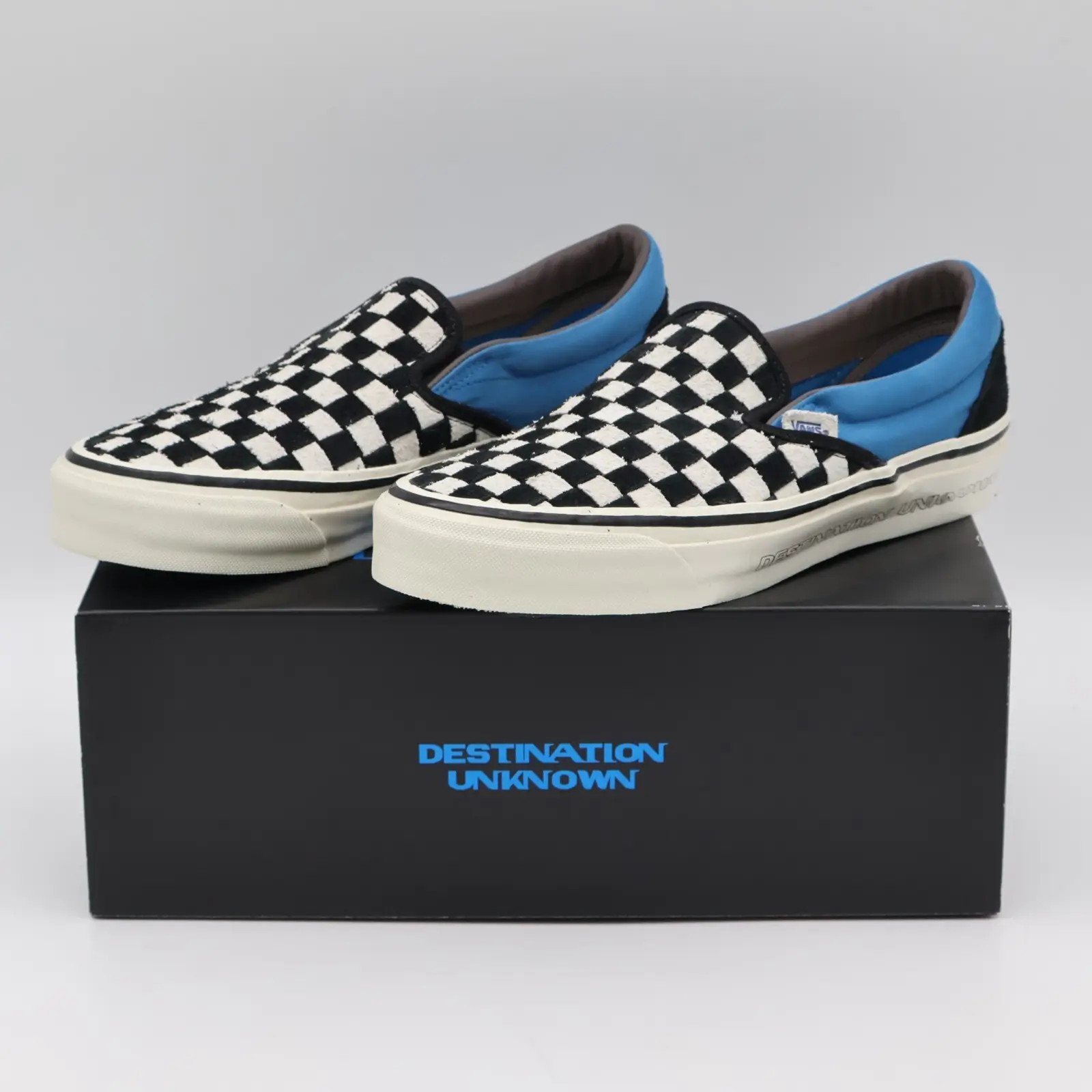 Pair of Vans x Destination Unknown Classic Slip-On sneakers resting on a black Destination Unknown shoebox. The shoes feature Vans' signature black-and-white checkerboard canvas upper, bright blue padded collars and lining, off-white foxing, and subtle Destination Unknown branding debossed along the midsole. The minimalist studio background highlights the collaborative details and vintage-inspired silhouette.