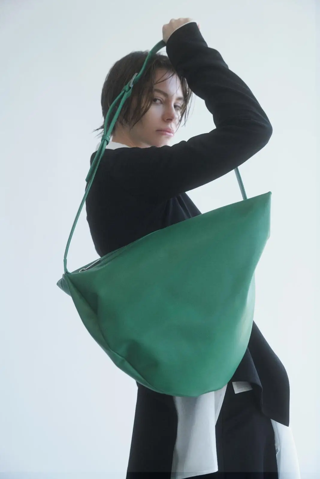 Model carrying a large emerald green leather shoulder tote with a wide adjustable strap, styled with a black long sleeve top against a clean white studio background.