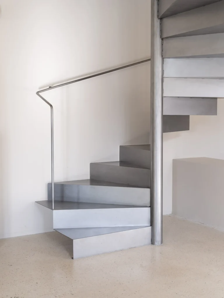 Industrial-style metal staircase with angular steel treads, slim stainless steel handrail, and polished concrete flooring in a bright minimalist interior