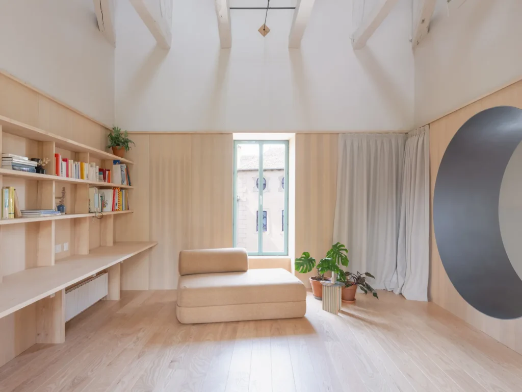Bright living space with exposed timber beams, built-in oak shelving, a minimalist lounge chair, indoor plants, and a large circular architectural open