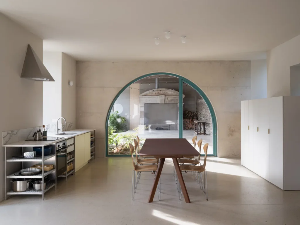 Open-plan dining room featuring a minimalist kitchen, large wooden dining table, concrete walls, and a dramatic arched glass opening overlooking a covered courtyard