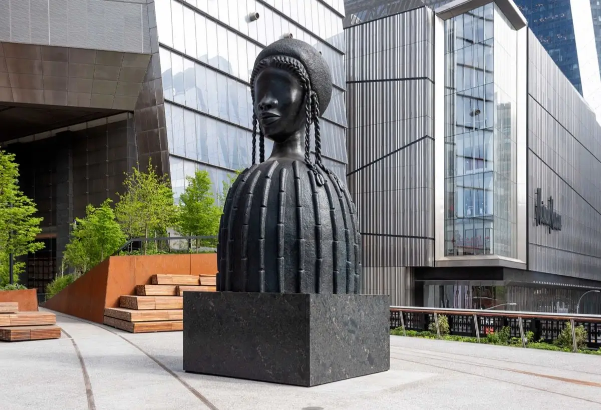 Simone Leigh's monumental bronze sculpture Brick House stands on the High Line Spur in Manhattan. The sculpture depicts a stylized Black woman with braided hair, a smooth eyeless face, and a cylindrical torso inspired by traditional African earthen architecture. Set atop a dark stone pedestal, the artwork is surrounded by landscaped seating and modern glass-and-steel buildings, emphasizing the dialogue between public art and contemporary urban architecture in New York City.