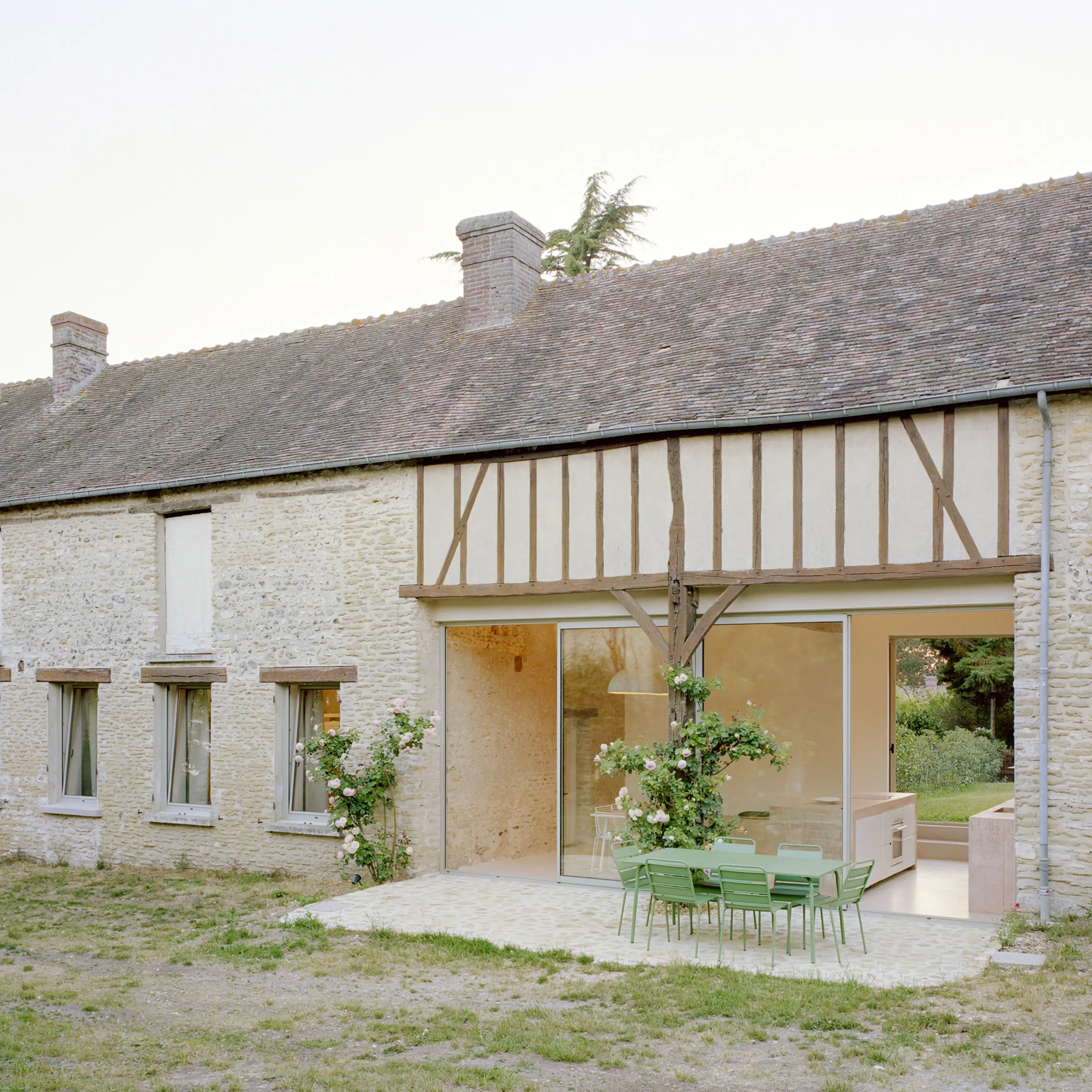 Renovated stone farmhouse with preserved timber framing, expansive sliding glass doors, and a contemporary patio furnished with green outdoor dining chairs overlooking a quiet garden.