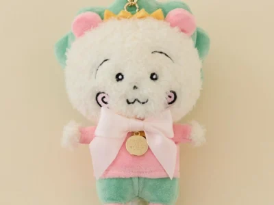 Plush COTI-COTI mascot keychain in pastel pink, mint green, and white with a satin bow, gold charm, and embroidered facial details, photographed against a cream background