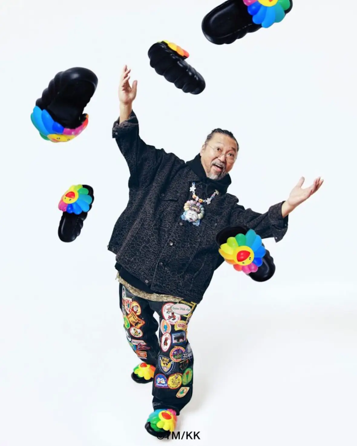 Takashi Murakami poses with arms raised as black sandals featuring his signature smiling rainbow flower appear to float around him against a white studio backdrop.