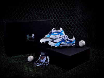 Two custom Under Armour baseball cleats in a blue-and-white space-inspired colorway displayed on a black presentation box beside a branded gift box, batting glove, and baseball against a dark backstop net background