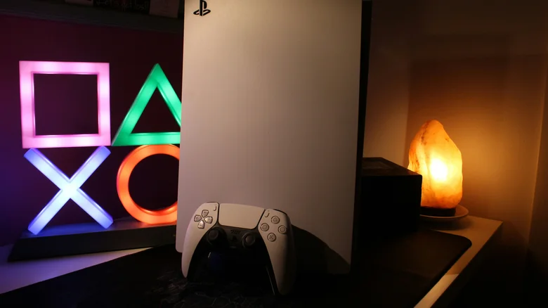 White PlayStation 5 console with a DualSense controller displayed beside illuminated PlayStation symbol lights and a warm glowing salt lamp on a modern gaming desk.