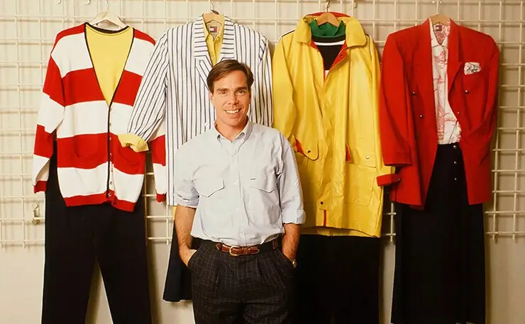 Vintage photograph of Tommy Hilfiger standing in front of colorful apparel samples, including striped knitwear, tailored jackets, and outerwear from the brand's early design years.