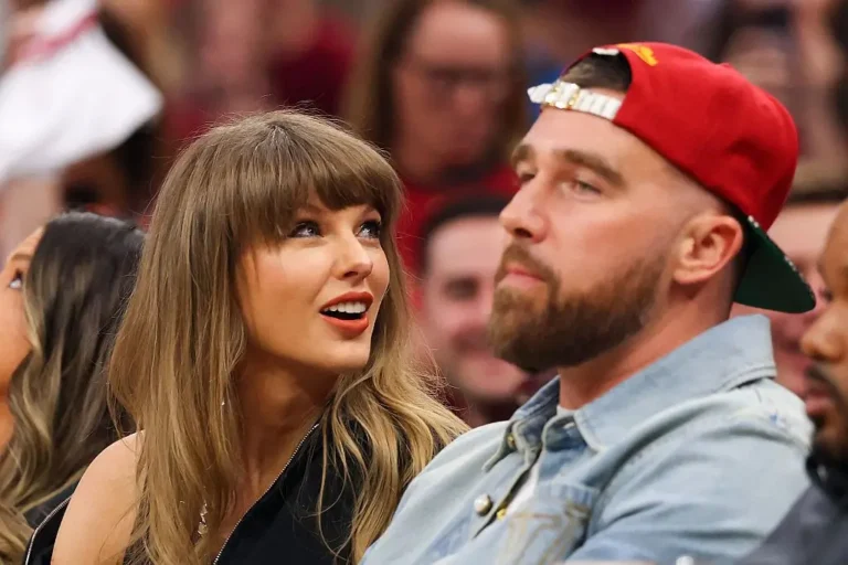 Taylor Swift and Travis Kelce sit courtside at a basketball game surrounded by a packed crowd. Swift wears a black sleeveless outfit with her signature bangs and looks toward Kelce while smiling mid-conversation. Kelce wears a light blue denim jacket and a red backward baseball cap, facing the court with a composed expression as fans fill the blurred arena background