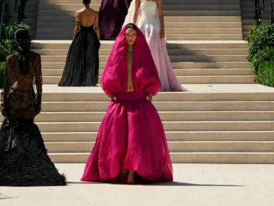 Fashion models present couture designs on a grand outdoor staircase framed by manicured greenery during a runway show. At center, a model wears a dramatic fuchsia hooded gown with sculptural volume and a deep front opening, while surrounding models showcase black lace, feathered, and ivory textured dresses, creating a striking contrast in color, sil and craft