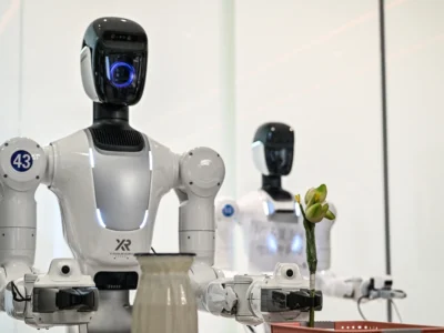 White service robot holding a small flower in a demonstration area, equipped with a large camera-like eye and articulated robotic arms. Additional robots stand behind it, highlighting hospitality and customer service applications in a clean exhibition set
