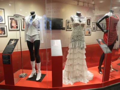 Museum display featuring iconic stage costumes, including a white sailor inspired outfit, an embellished white ruffled gown, and a sparkling red dress on mannequins behind glass, with framed photos and exhibit panels in the background