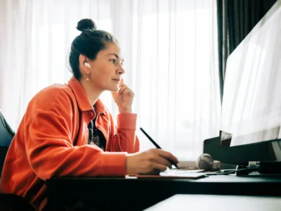 Person in an orange jacket and wireless earbuds works at a desktop computer with a pen tablet and stylus beside the keyboard. Sunlight filters through sheer curtains behind the workstation, illuminating a desk that also holds over-ear headphones and other creative work accessor