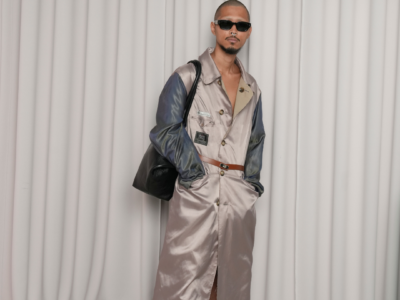 Full-length portrait of a model wearing a glossy taupe satin trench-style coat with iridescent blue sleeves, cinched with a slim brown belt. The look is styled with black sunglasses, a large black shoulder bag, and oversized plush animal slippers, standing in front of soft white draped curtains for a contemporary fashion presentation