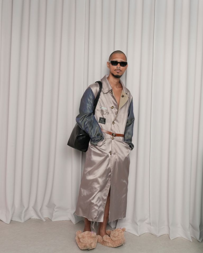 Full-length portrait of a model wearing a glossy taupe satin trench-style coat with iridescent blue sleeves, cinched with a slim brown belt. The look is styled with black sunglasses, a large black shoulder bag, and oversized plush animal slippers, standing in front of soft white draped curtains for a contemporary fashion presentation
