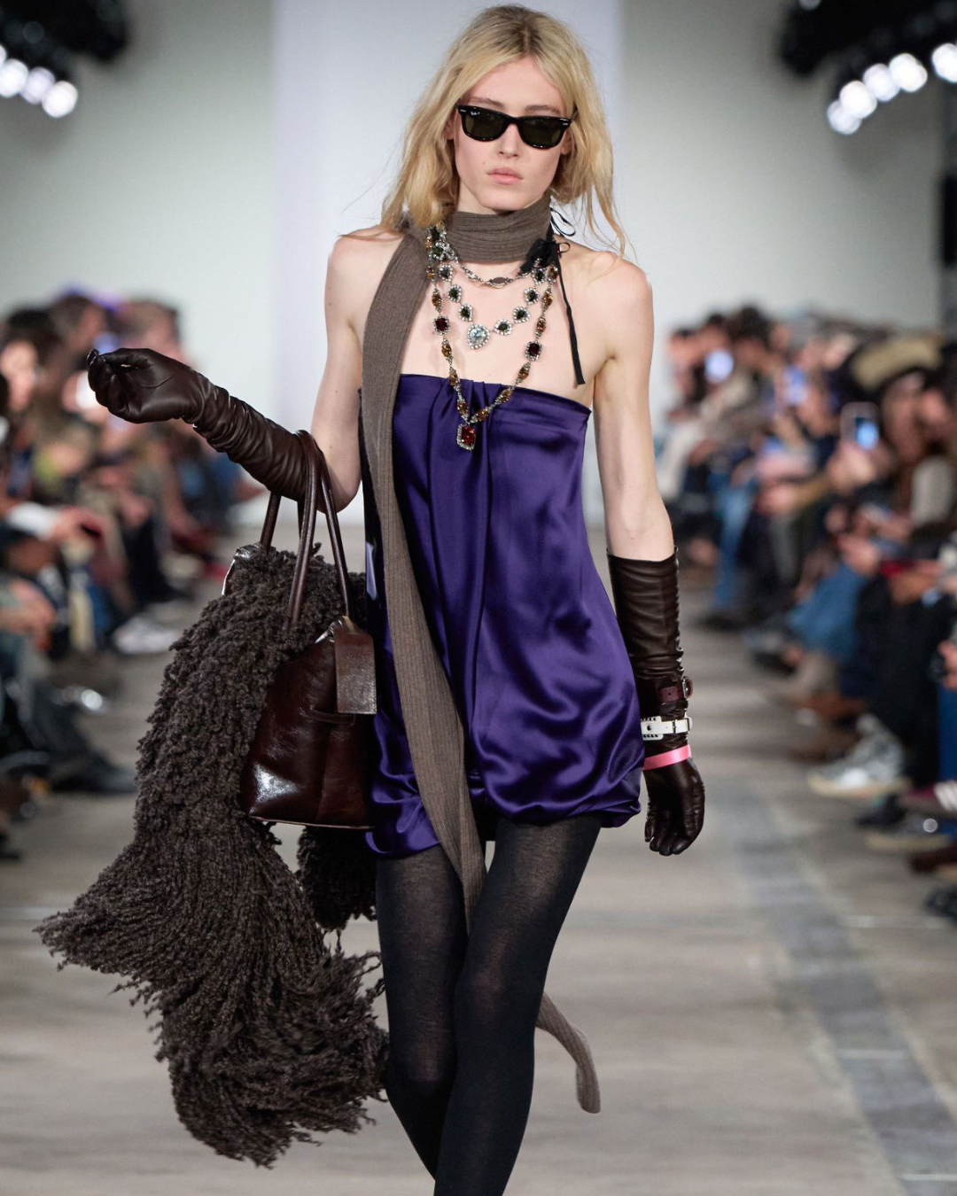 Runway model wearing a deep purple satin mini dress with layered statement necklaces, a long taupe scarf, black leather gloves, sheer black tights, dark sunglasses, and carrying a large brown leather handbag with a textured knit wrap during a luxury fashion show.