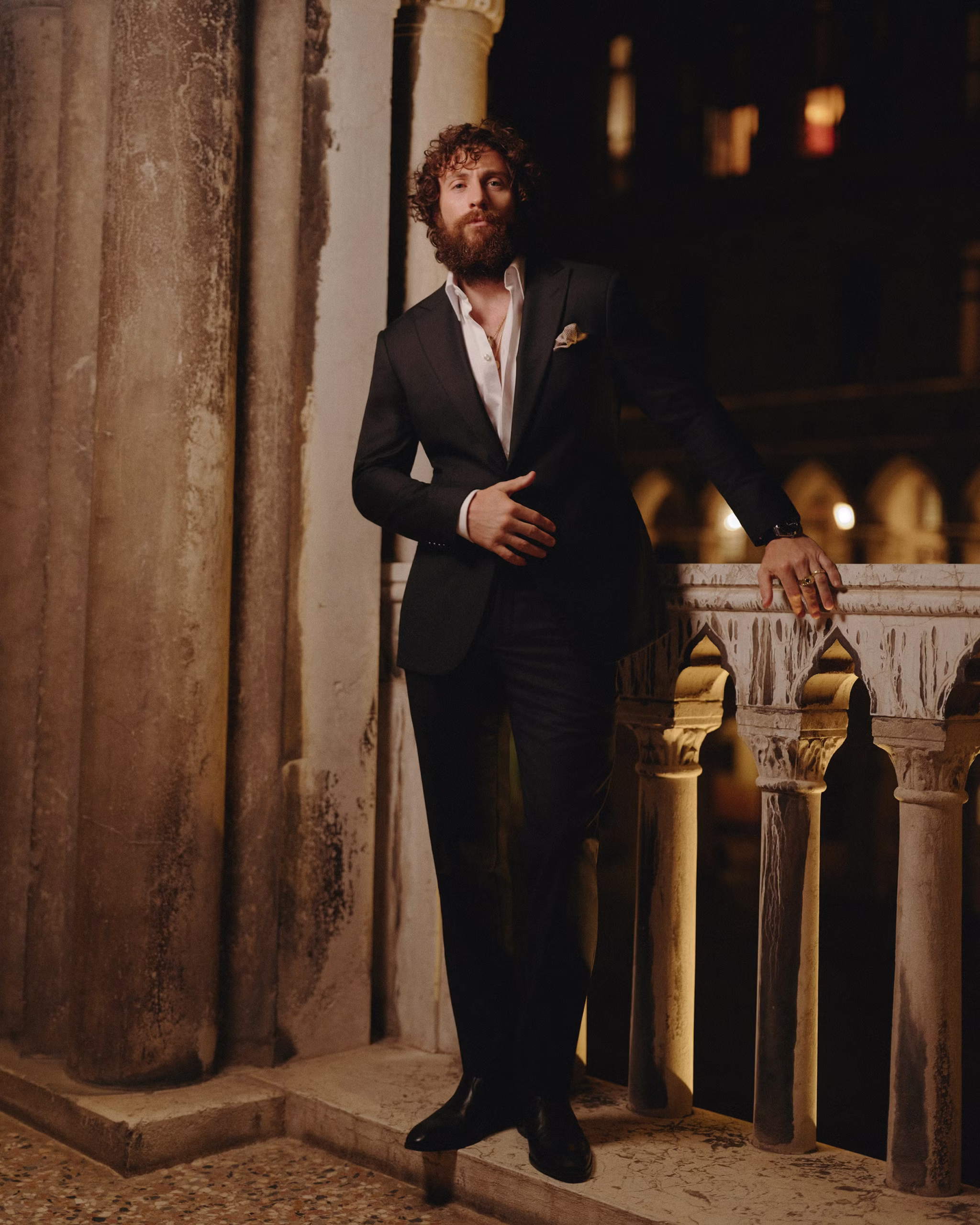 Man wearing a tailored black suit with an open-collar white dress shirt stands beside an ornate stone balustrade in a historic courtyard at night, illuminated by warm ambient lighting while wearing a dark luxury wristwatch.
