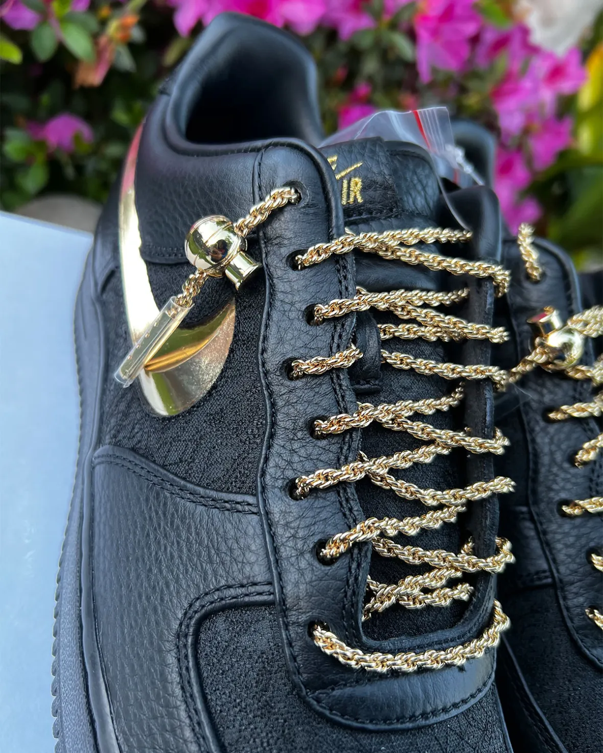 Close-up of the Victor Solomon x Nike Air Force 1 in black premium leather, highlighting its metallic gold chain laces, polished gold Swoosh, luxury-inspired lace hardware, and textured materials against a vibrant floral background.
