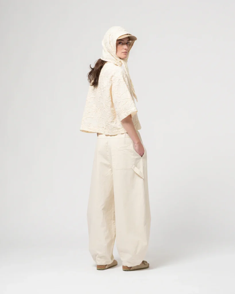 Model wearing a textured cream hooded cape layered over wide-leg cream trousers, styled with a matching cap and casual sandals for a monochrome look