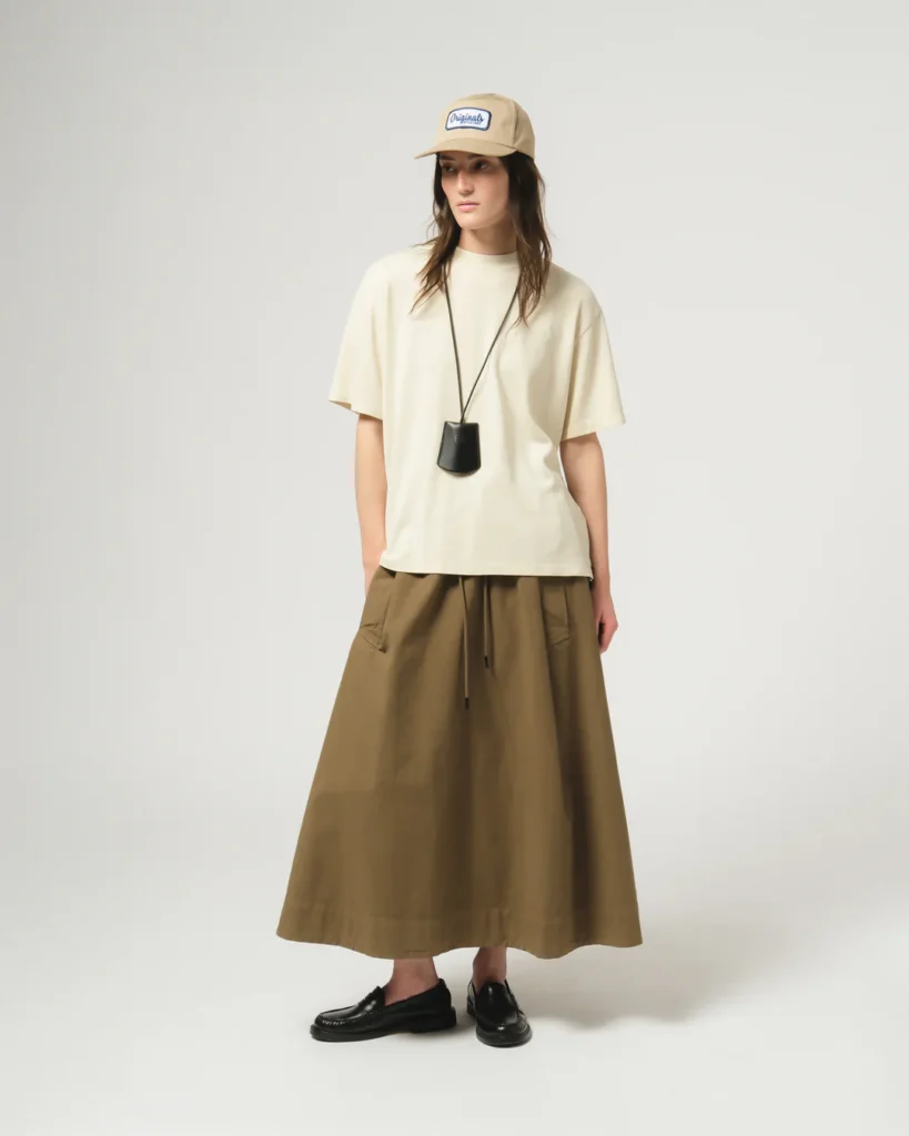 Model wearing an oversized cream T-shirt with a khaki drawstring midi skirt, beige logo cap, black leather pouch necklace, and black loafer