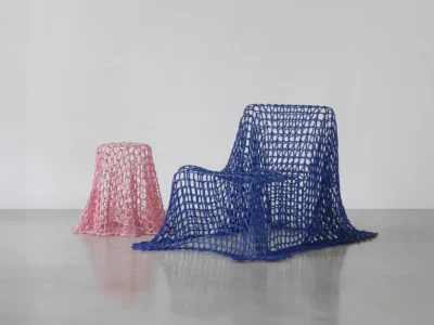 Colorful woven furniture collection: A deep blue woven lounge chair and a pastel pink woven stool, both draped in sculptural open-mesh textile forms, displayed together in a minimalist gallery with polished concrete floor