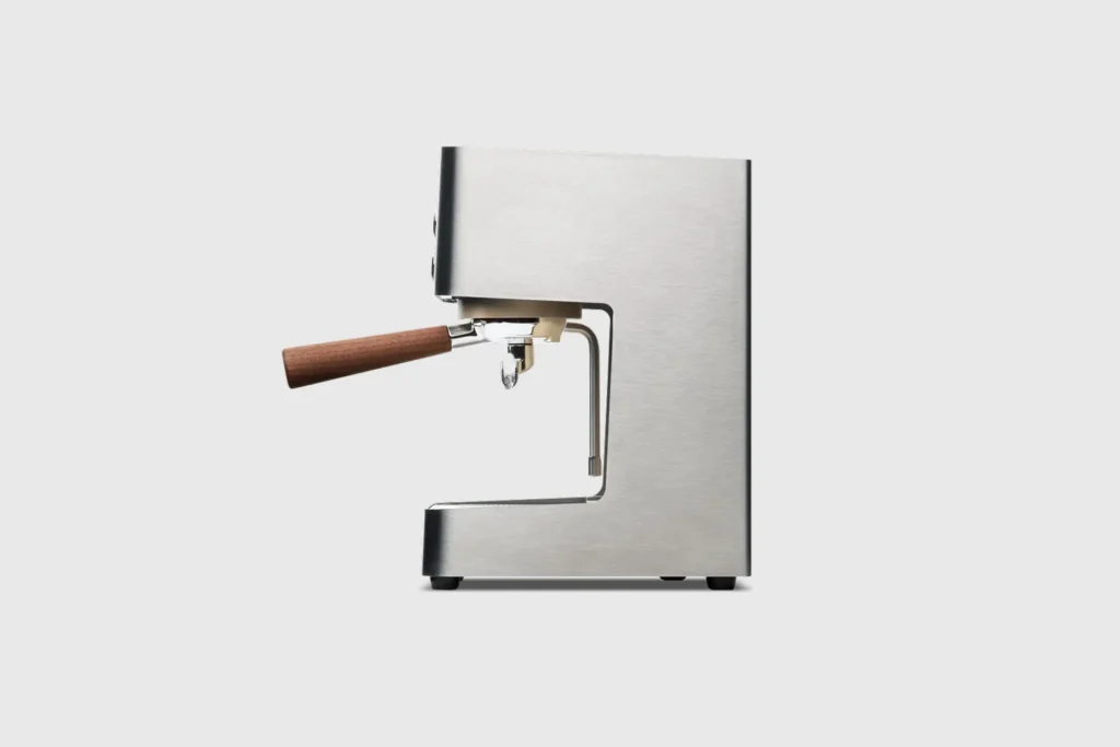 Side profile of a compact stainless steel espresso machine featuring a minimalist design, wooden portafilter handle, and integrated steam wand on a clean studio background