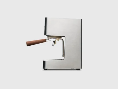Side profile of a compact stainless steel espresso machine featuring a minimalist design, wooden portafilter handle, and integrated steam wand on a clean studio background