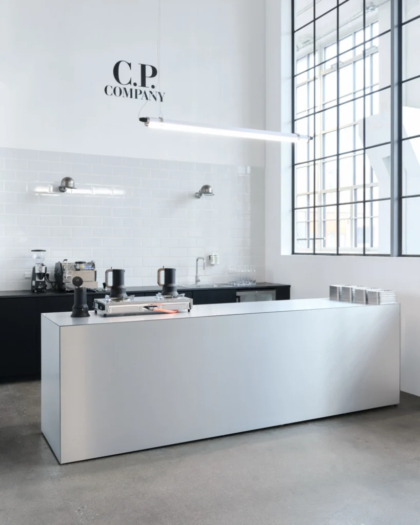Min C.P. Company exhibition café with a brushed-metal counter, coffee equipment, white tiled wall and large industrial window