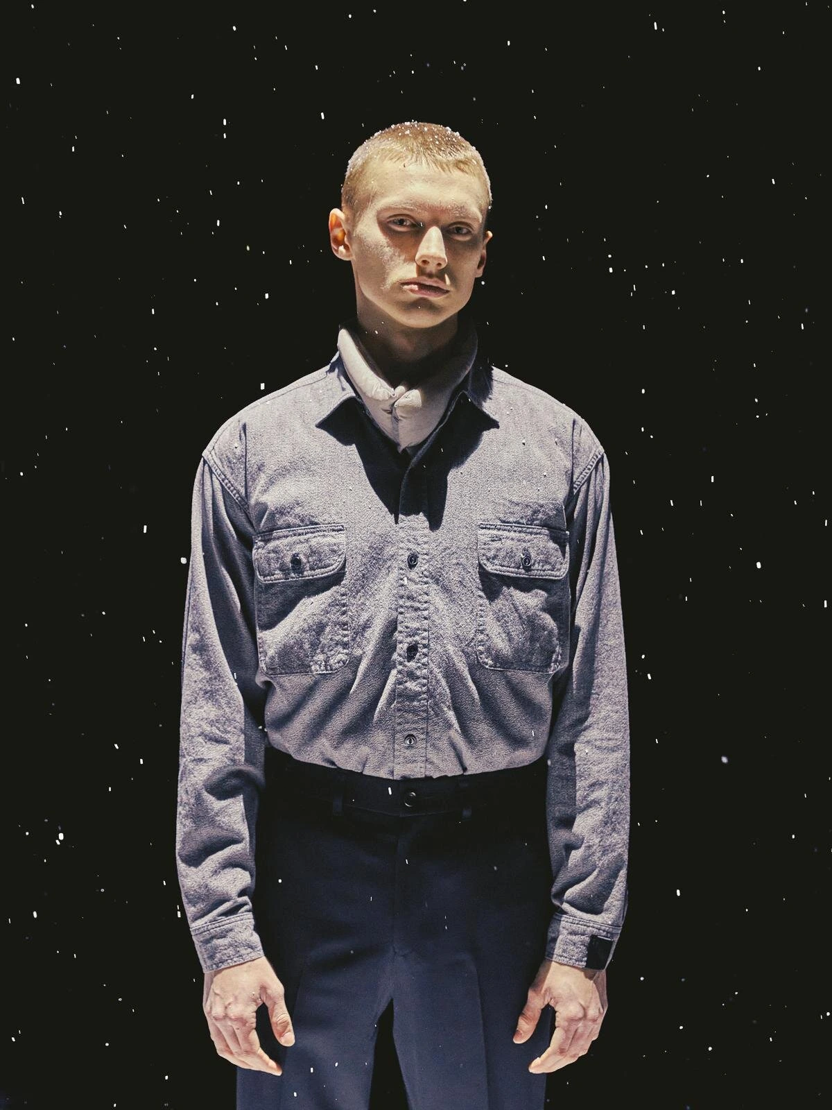 Model wearing a grey work shirt with dual chest pockets and dark trousers under dramatic lighting, surrounded by falling white particles against a black background.