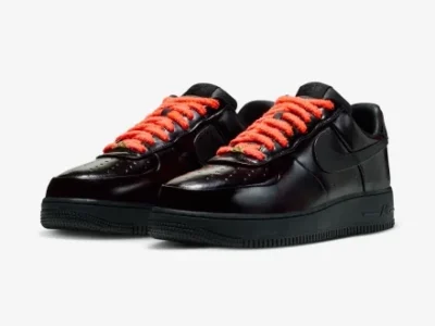 A 3/4 front angle of a pair of black patent leather Nike Air Force 1 sneakers with bright orange fuzzy laces, showcasing the glossy toe box and side profile