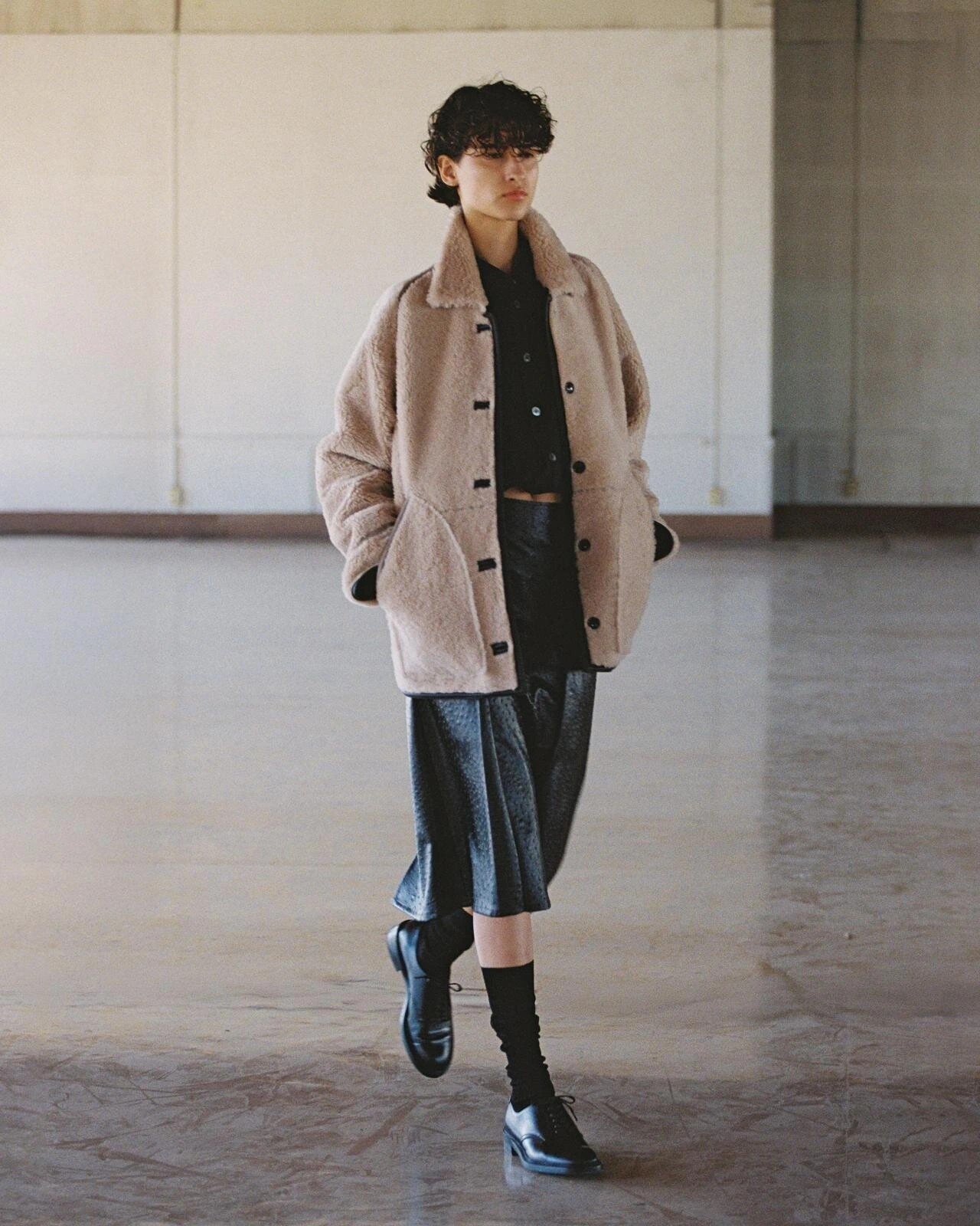 Model poses in a beige shearling jacket, black shirt and cropped leather trousers in a spacious interior