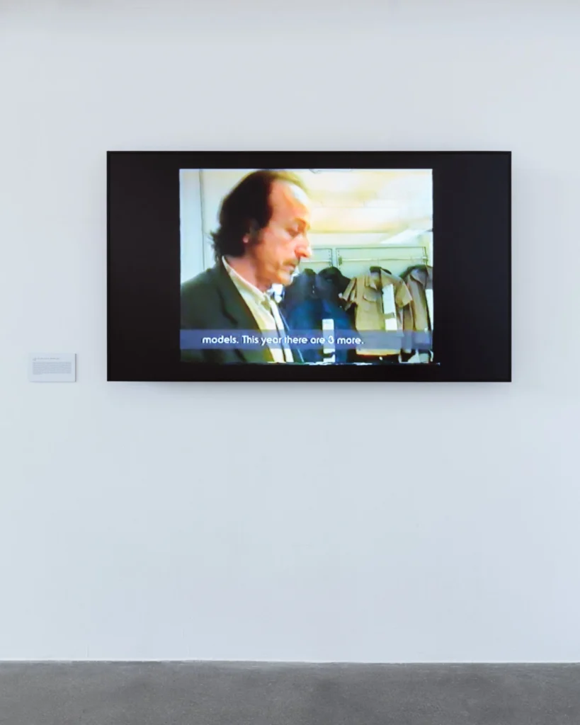 Wall-mounted screen showing archival C.P. Company footage of a man speaking beside hanging jacket