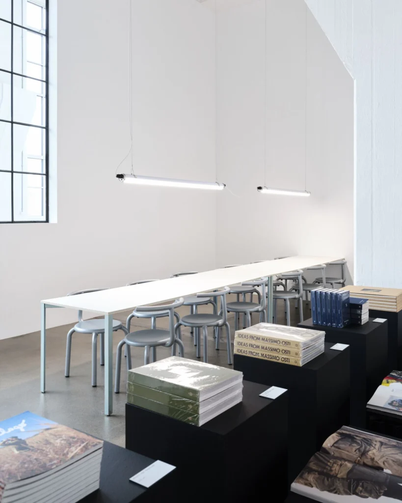 Exhibition reading area with long white tables, gray chairs, suspended tube lights and stacks of C.P. Company books and pub