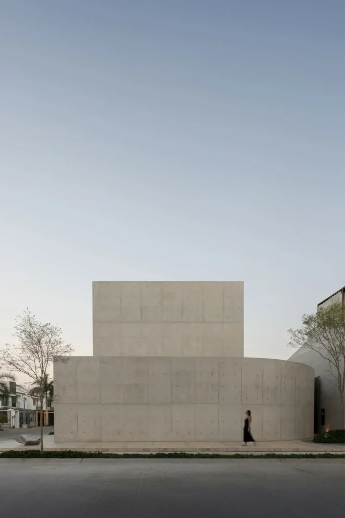 Modern min concrete residence with a curved exterior wall, clean geometric forms, and a solitary figure walking along the sidewalk beneath a clear blue sky