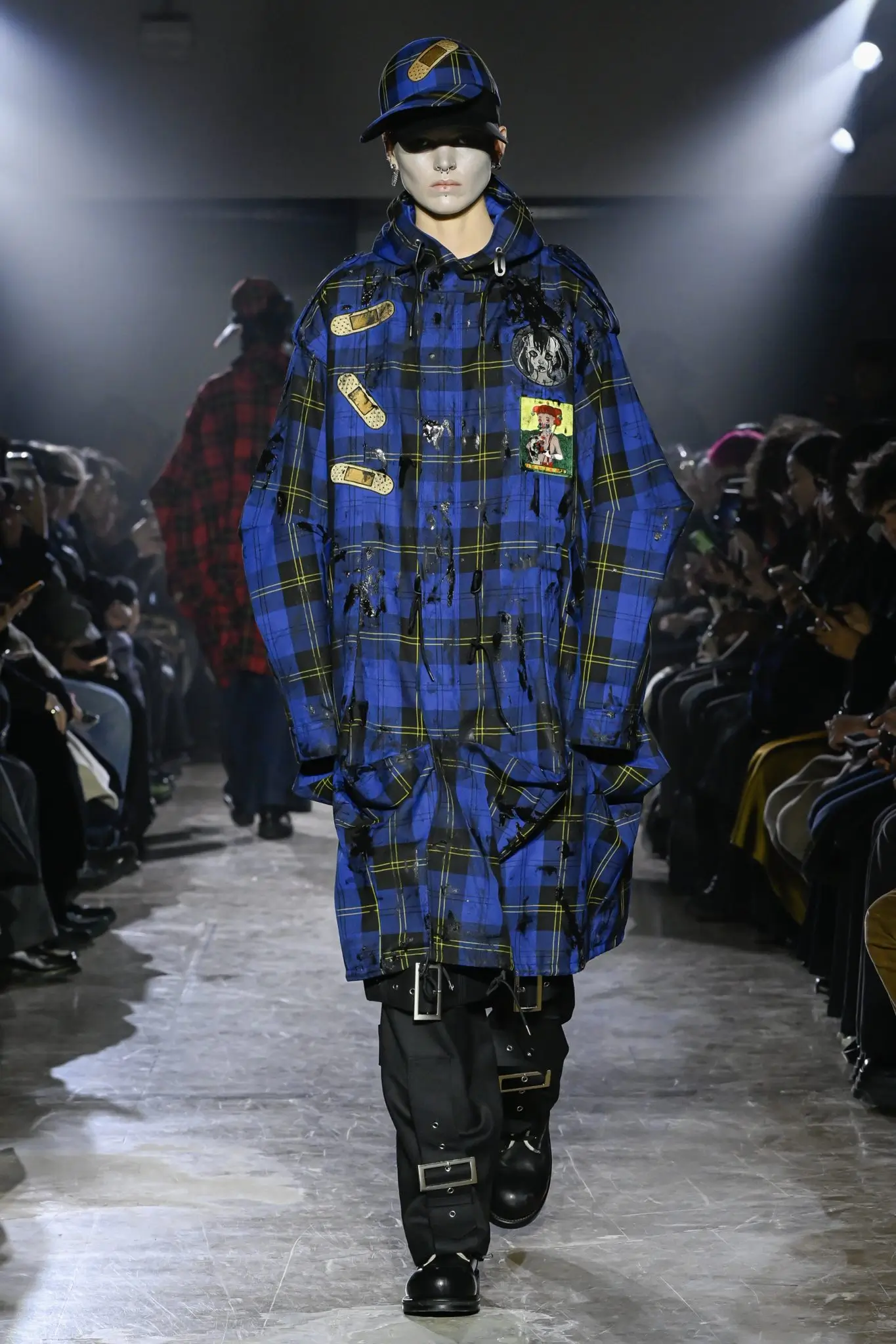 Model On The Runway Wearing An Oversized Blue Tartan Hooded Coat With Graphic Patches, Black Buckled Trousers And Matching Tartan Cap.