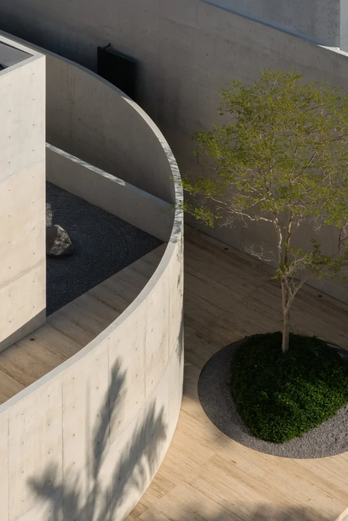 Elevate view of a sculpture curved concrete courtyard wall surround a Zen-inspired gravel garden, mature tree, and warm stone pave