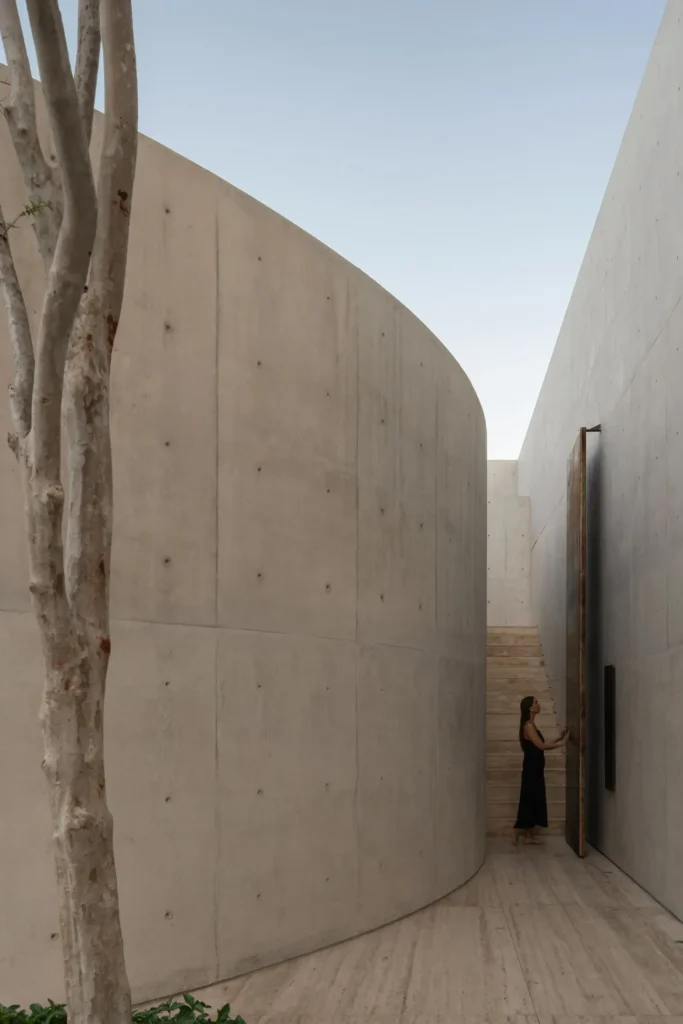 Narrow outdoor passage frame by smooth expose concrete walls, a tall pivot door, staircase, and a person enter the tranquil mi home