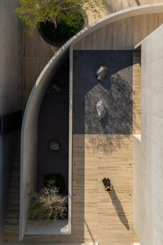 Overhead view of a min courtyard feature curved concrete walls, raked gravel, natural stone elements, warm stone flooring, and a lone figure cast a long sil