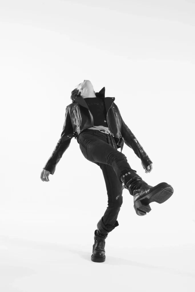 Model balancing mid-stride with head thrown back, wearing a cropped black leather biker jacket, slim black jeans, and stacked leather boots in a minimalist white studio