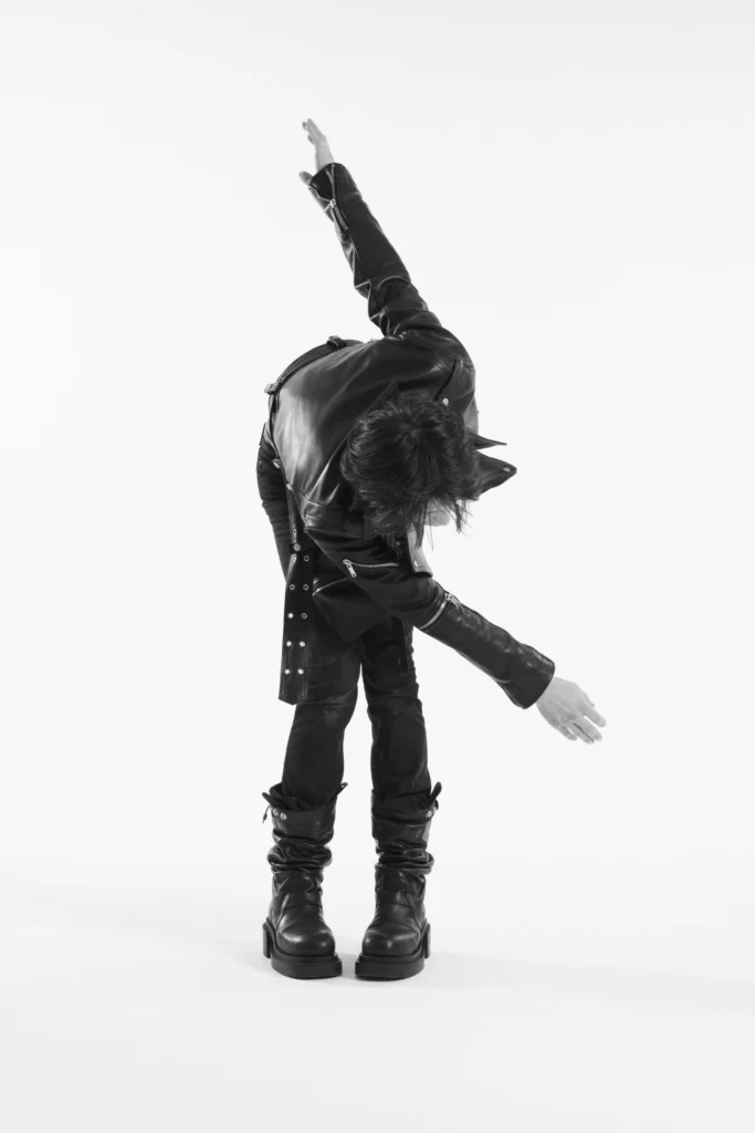 Monochrome studio photograph of a model bending sideways with outstretched arms, showcasing a black leather biker jacket, fitted denim, and slouchy leather boot