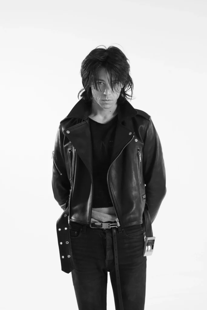 Front-facing portrait of a model in a cropped black leather jacket, black knit top, slim jeans, and a long hanging belt against a clean white background