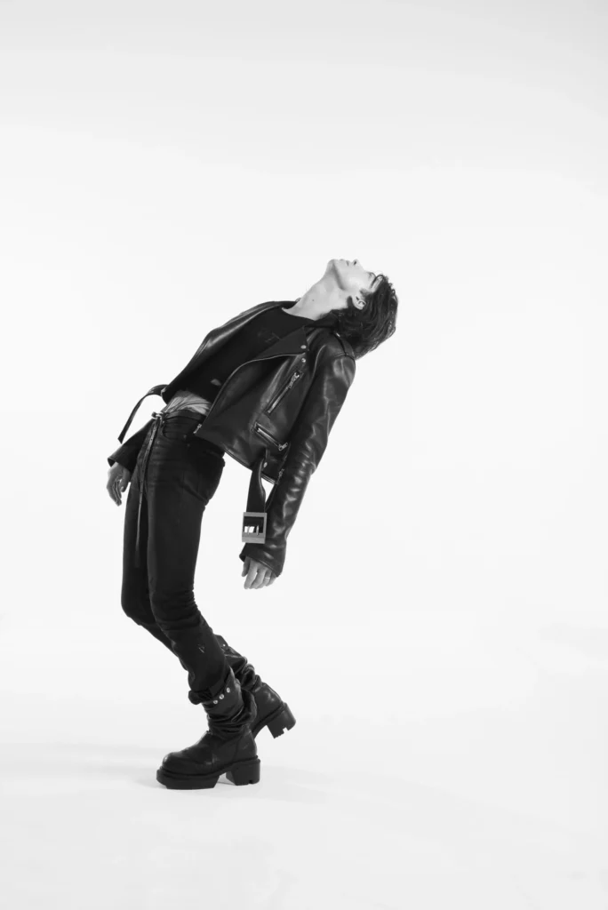 Side-profile fashion portrait of a model leaning dramatically backward in a cropped leather jacket, slim black jeans, and heeled leather boot