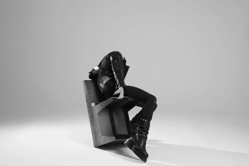 Minimalist monochrome image of a model reclining across an angular sculptural chair, emphasizing sharp geometry and tailor leather style