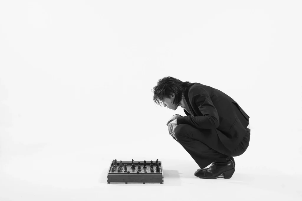 Black-and-white studio portrait of a crouching model studying a handcrafted chessboard, dressed in a tailored black jacket, trousers, and heeled boot