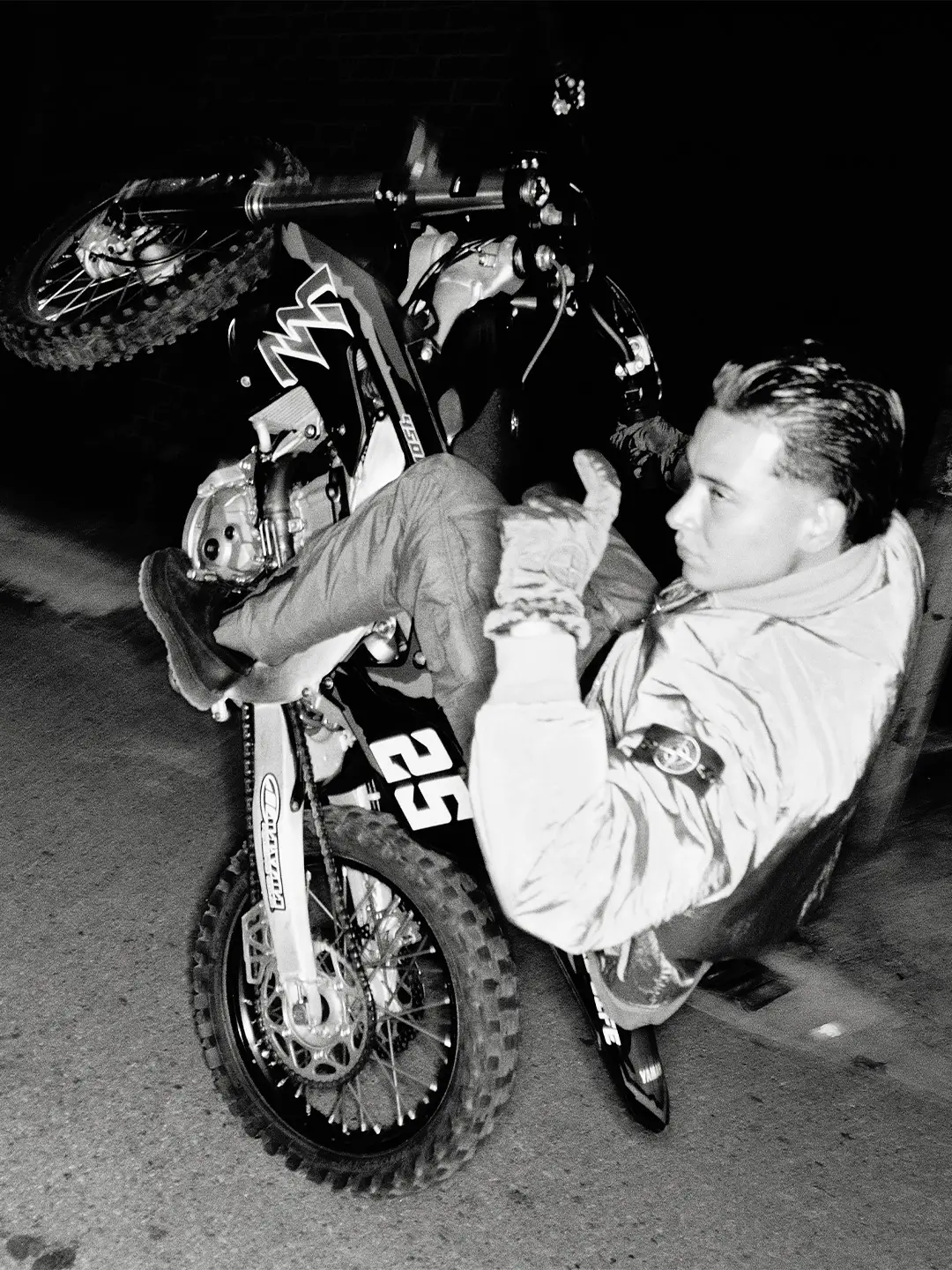 Black-and-white nighttime photograph of a person in a Stone Island jacket reclining against a dirt bike while holding a small cat, captured with stark flash photography.