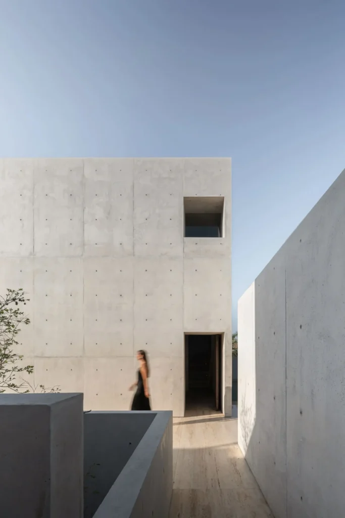Sunlit concrete residence with monolithic architecture, narrow walkways, recess entry, and a blurred figure move through the min outdoor space