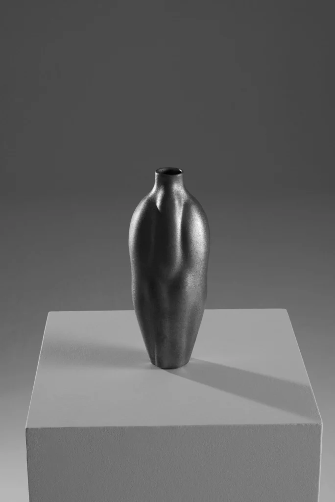 Minimalist studio photograph of a sculpture matte black ceramic vase with a softly contoured sil display on a white pedestal