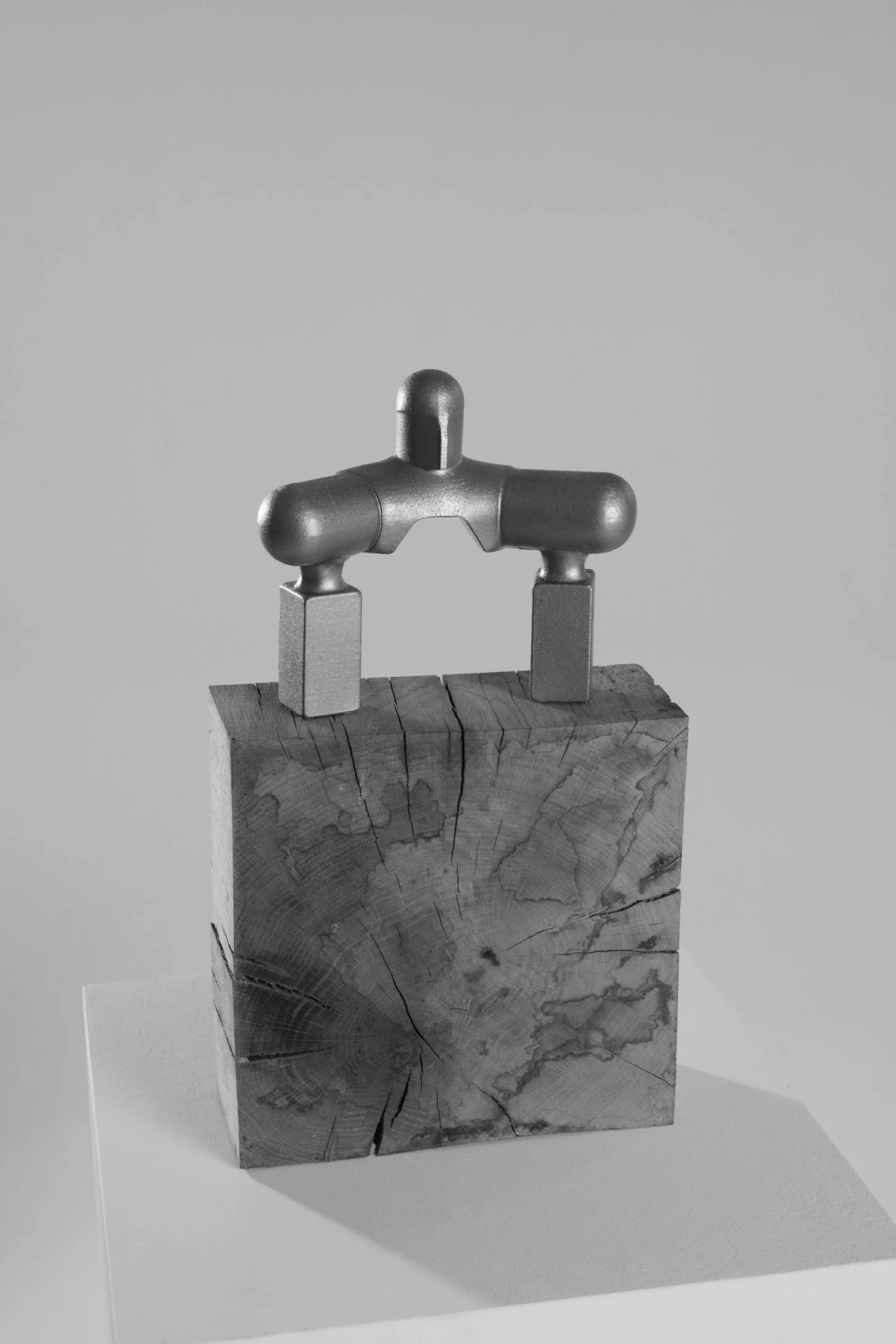 Minimalist metallic sculpture with rounded geometric forms balanced on two rectangular supports atop a weathered solid wood block, displayed against a clean neutral backdrop.