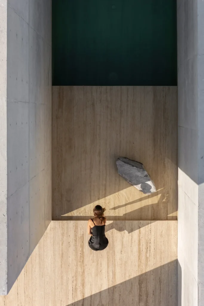 Top-down architectural view of a serene reflecting pool, floating stone, and seated figure enclosed by tall exposed concrete walls with dramatic afternoon light